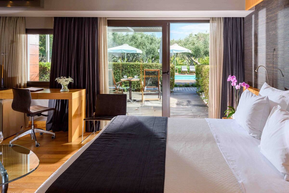 Une chambre d’hôtel avec un lit, un bureau et une chaise, une table en verre avec une bouteille, et une vue sur l’espace extérieur de la piscine.