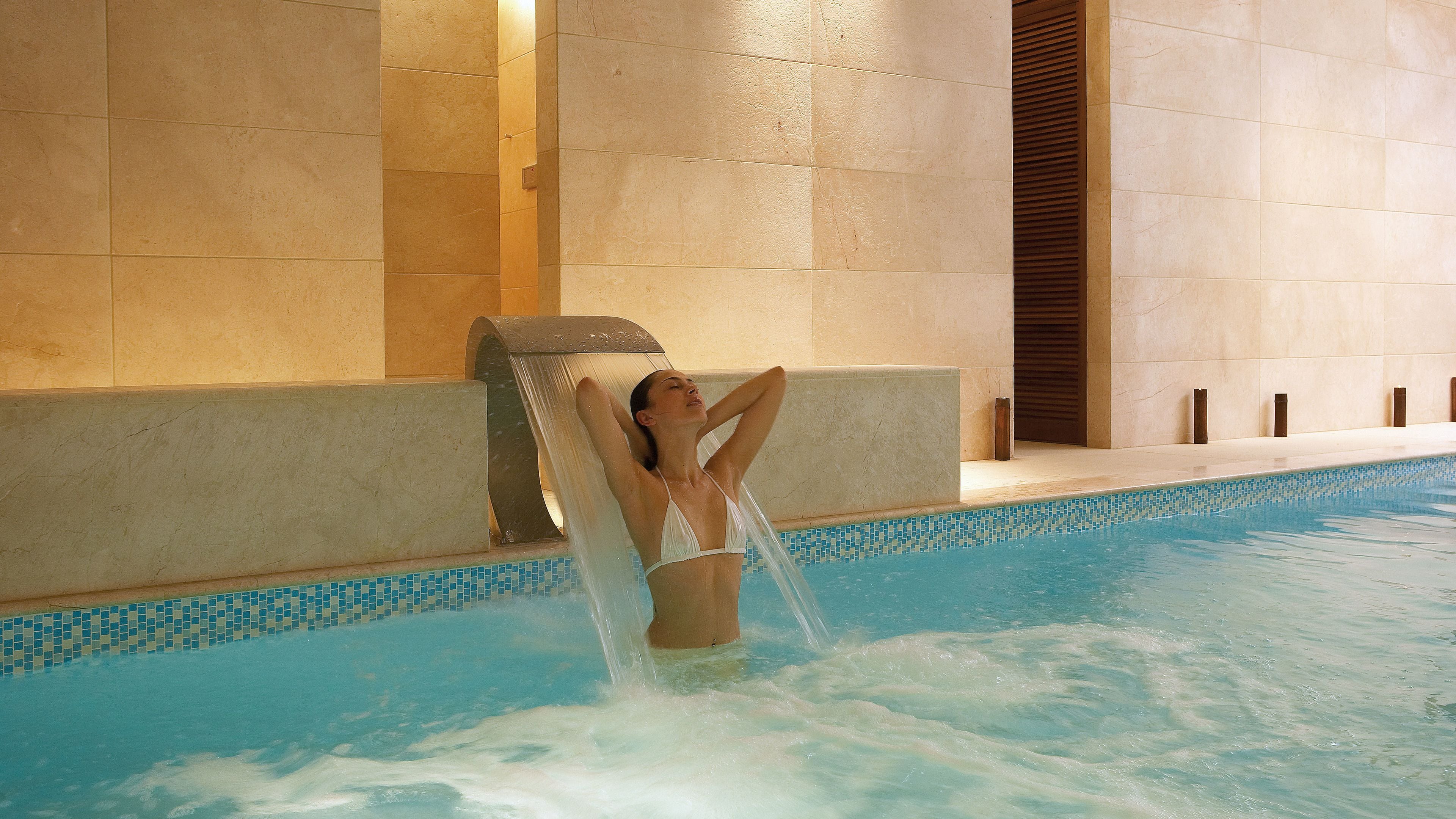 A woman relaxing in a pool with a water feature, surrounded by tiled walls.