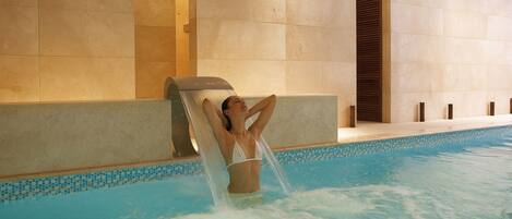 A woman relaxing in a pool with a water feature, surrounded by tiled walls.
