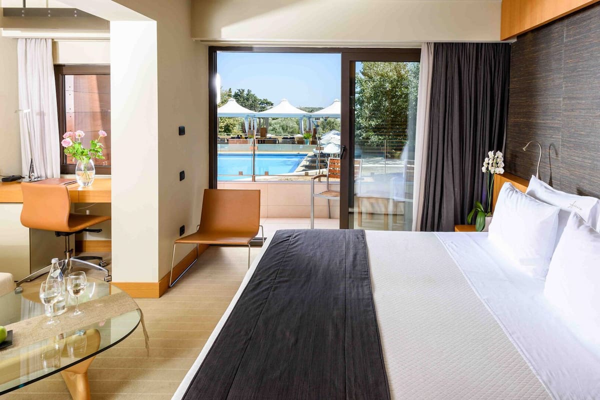 Une chambre d’hôtel avec un grand lit, un bureau avec une chaise, une table en verre avec deux verres à vin et une pomme, et une vue sur une piscine et des parasols à l’extérieur.