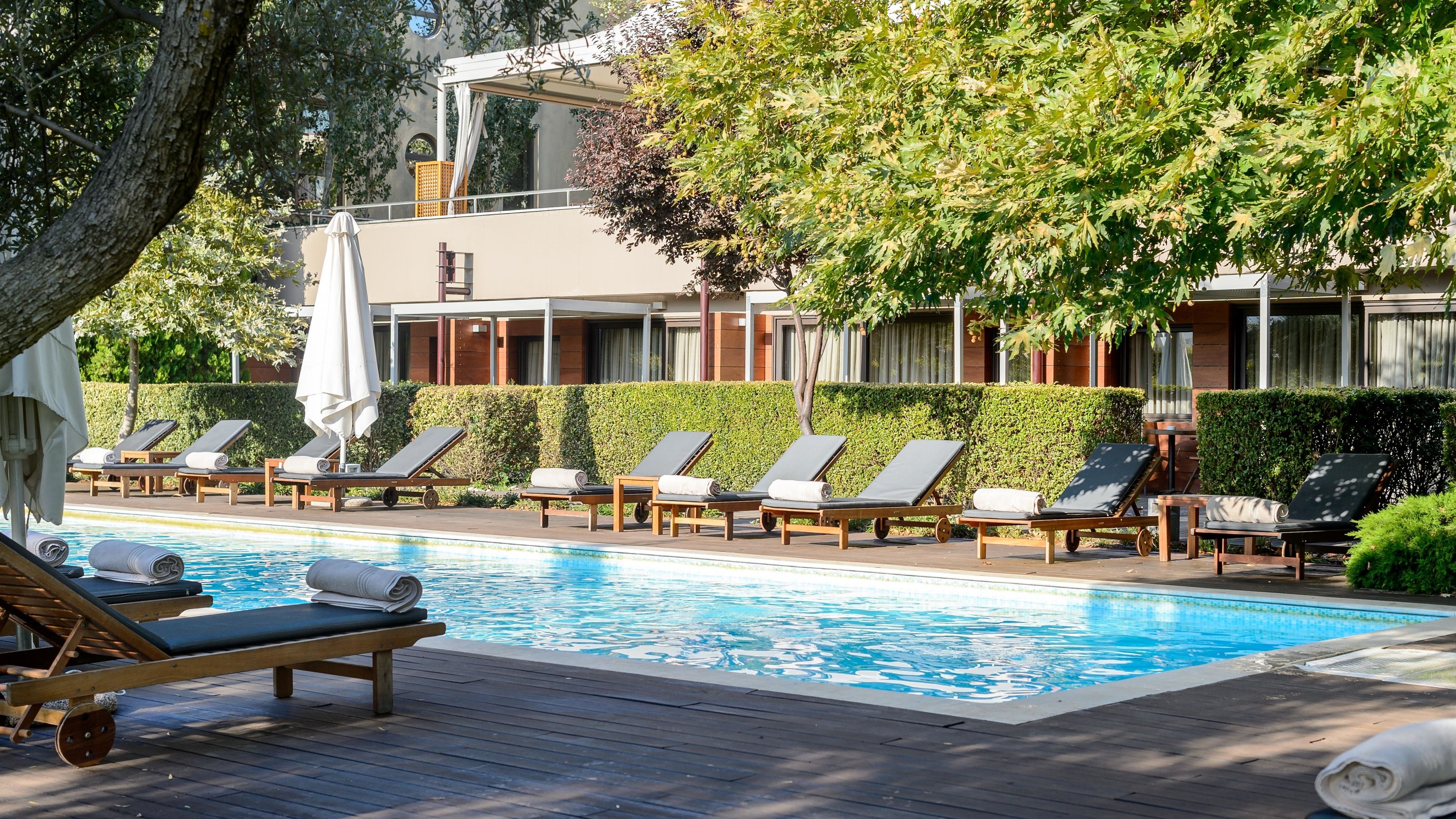 Un espace aménagé au bord de la piscine, avec des chaises longues, une terrasse en bois et un bâtiment doté de grandes fenêtres, le tout agrémenté d’une haie en arrière-plan.