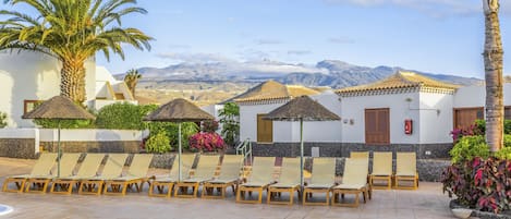 A resort with sun loungers, palm trees, and a mountain view.