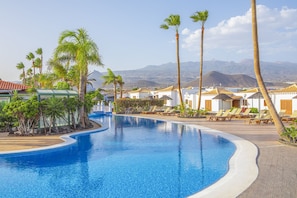A pool with a curved edge, surrounded by palm trees and lounge chairs, with mountains in the background.