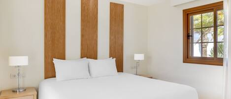 Habitación de hotel ordenada con una cama grande, dos mesitas de noche con lámparas, un cabecero de madera y una ventana con cortinas.
