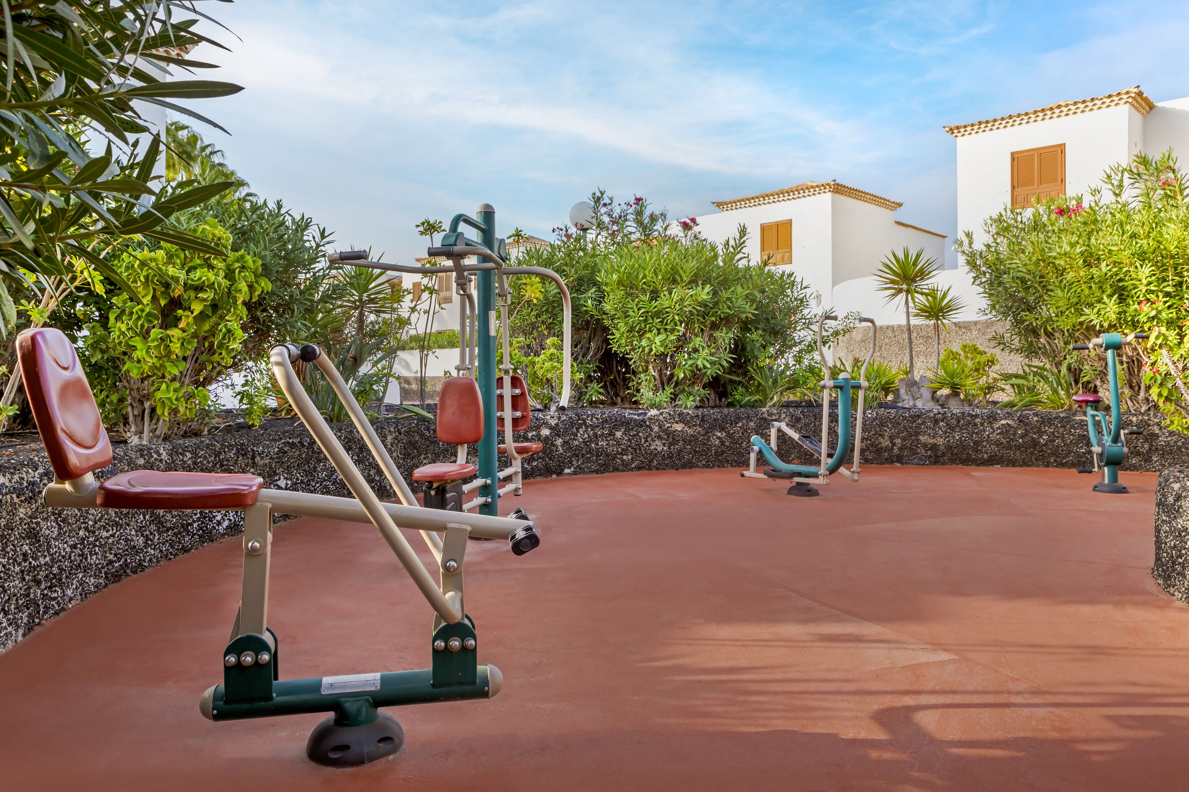 An outdoor gym area with exercise equipment, surrounded by greenery and a building in the background.