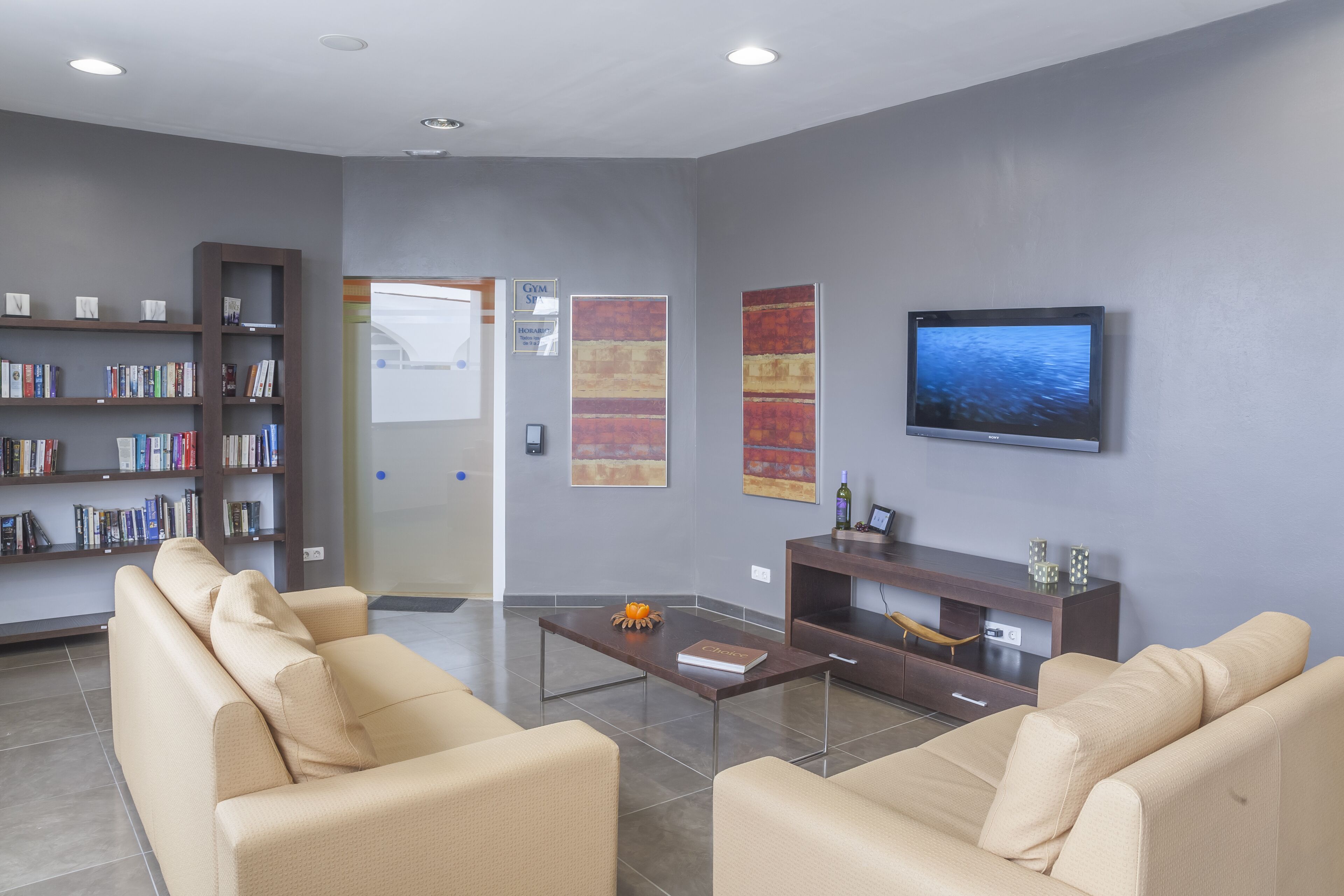 A modern lobby with beige sofas, a coffee table, a bookshelf, a flat-screen TV, and abstract art on the walls.