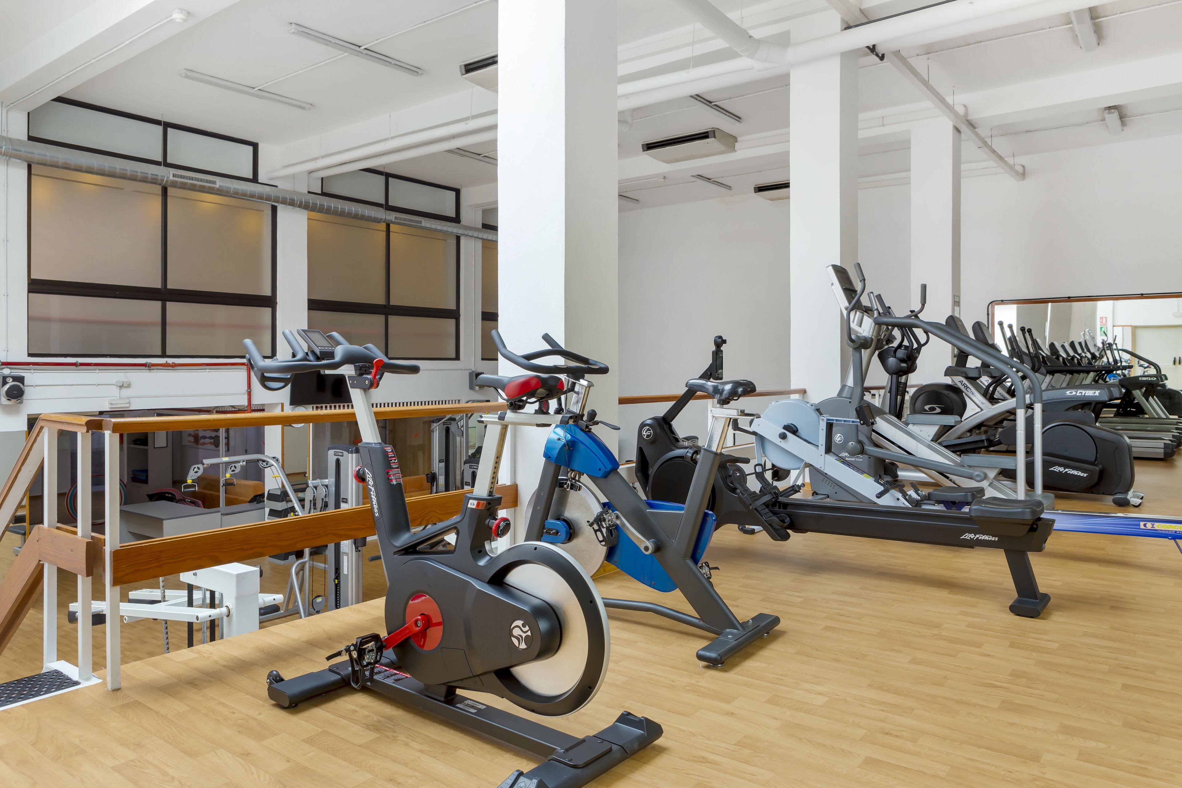 A gym with various exercise machines, including stationary bikes and treadmills, arranged on a wooden floor.