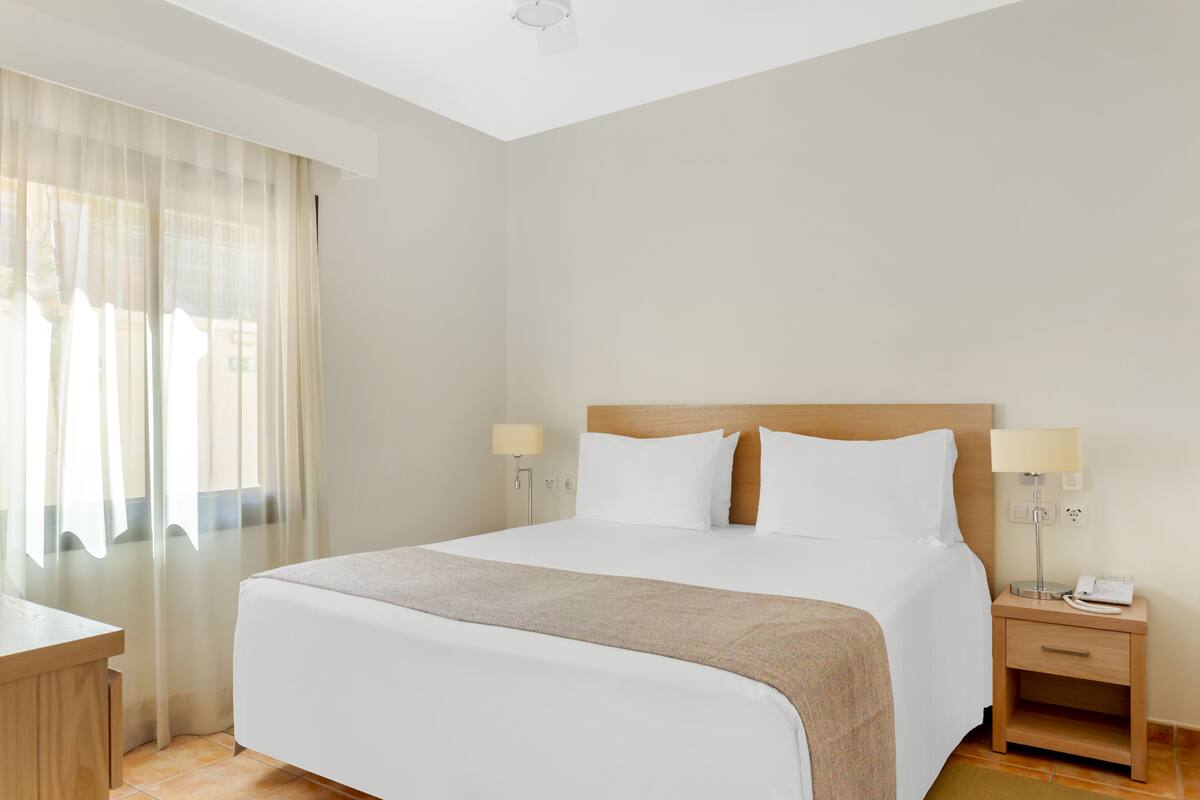 Quarto de hotel com uma cama grande, duas mesas de cabeceira com abajures, uma cabeceira de madeira e cortinas leves.