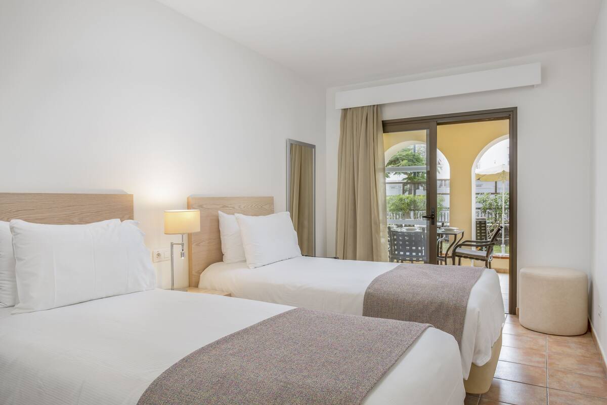 Quarto de hotel com duas camas, uma varanda com vista e uma pequena mesa e cadeira.