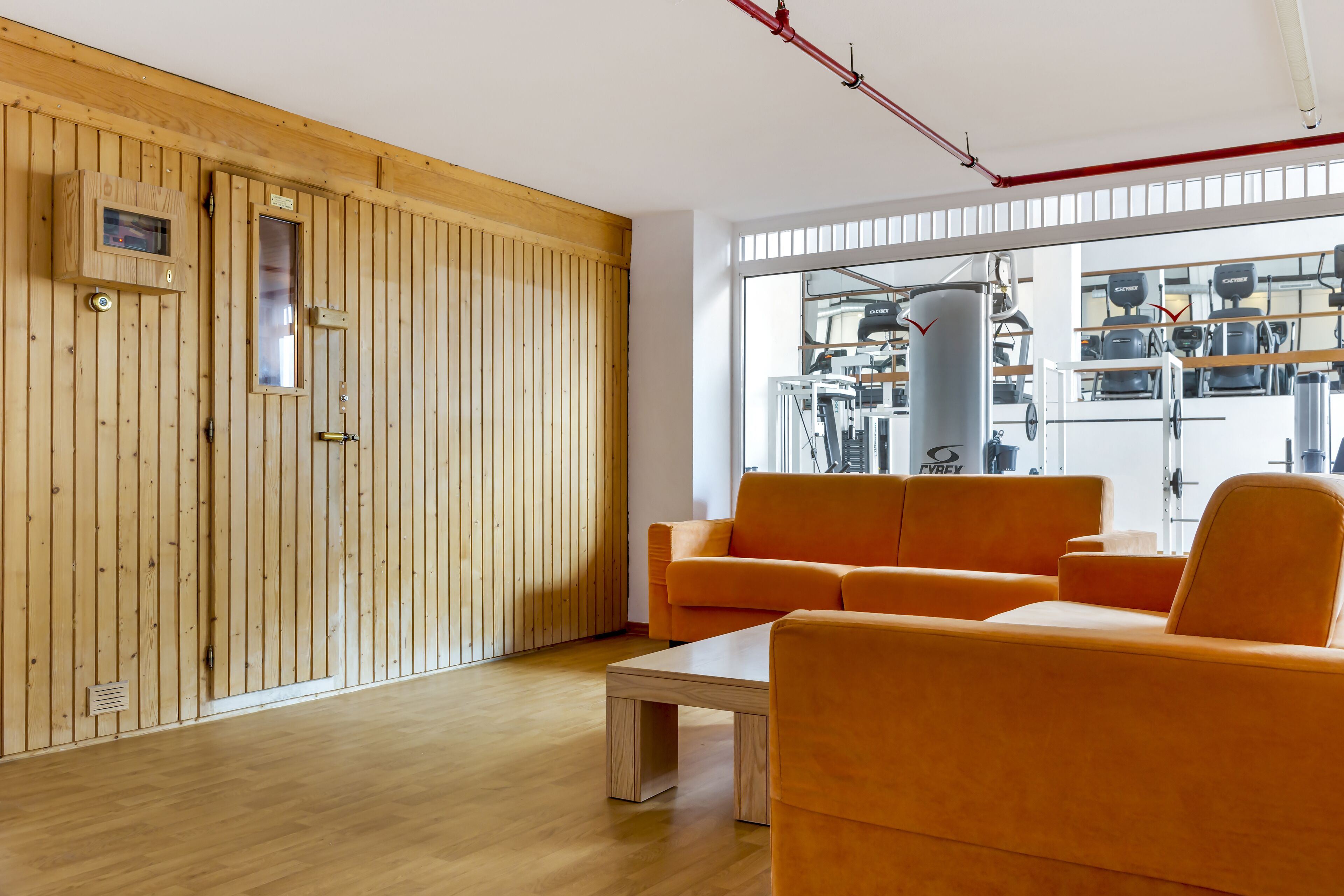 Een kamer met houten paneelwanden, een groot raam met zicht op fitnessapparatuur, en een oranje bank.