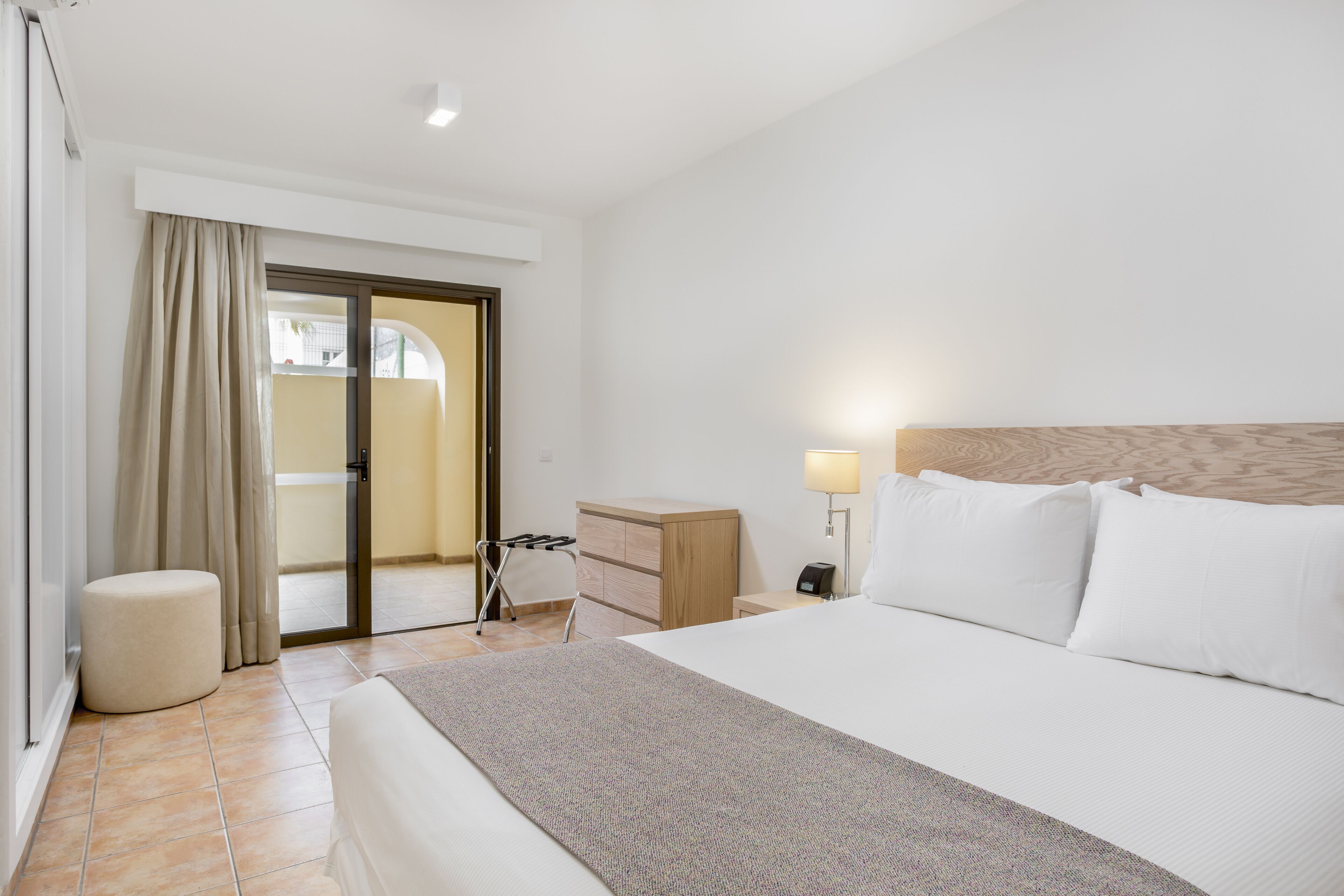 A hotel room with a bed, a bedside table, a lamp, a dresser, a chair, and a sliding door leading to an outdoor area.