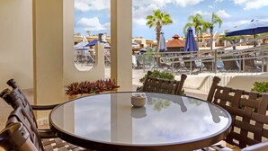 Une table à plateau en verre, avec des chaises et un petit bol, donnant sur un espace piscine agrémenté de parasols et de palmiers.