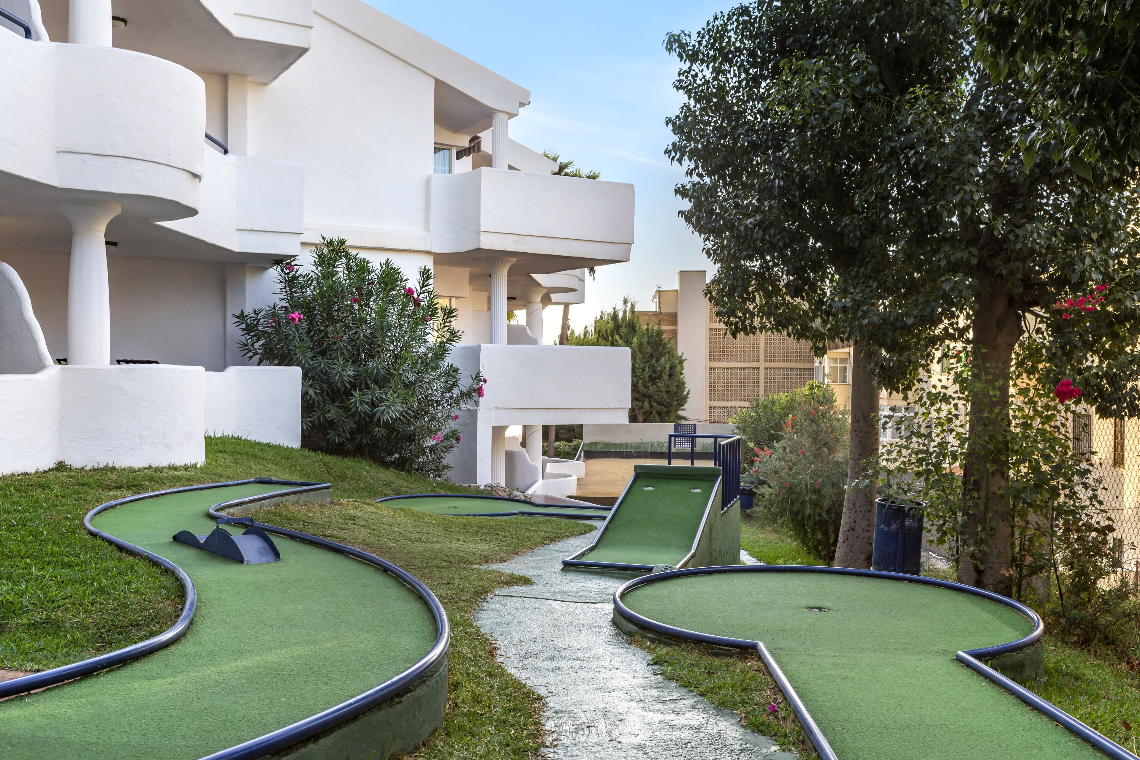Un campo de mini golf con un sendero sinuoso, rodeado por un edificio blanco con balcones y vegetación.