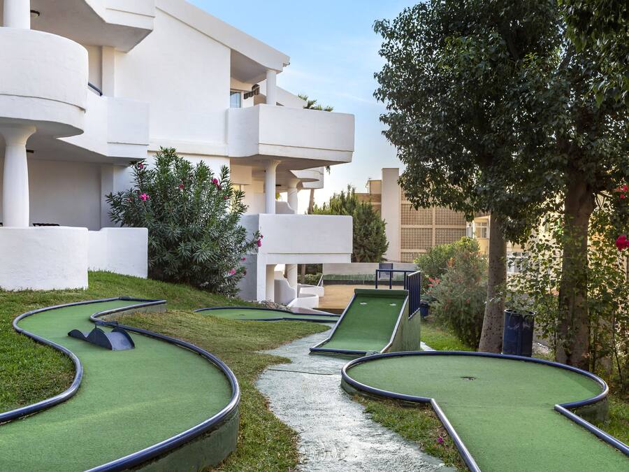 Un campo de mini golf con un sendero sinuoso, rodeado por un edificio blanco con balcones y vegetación.