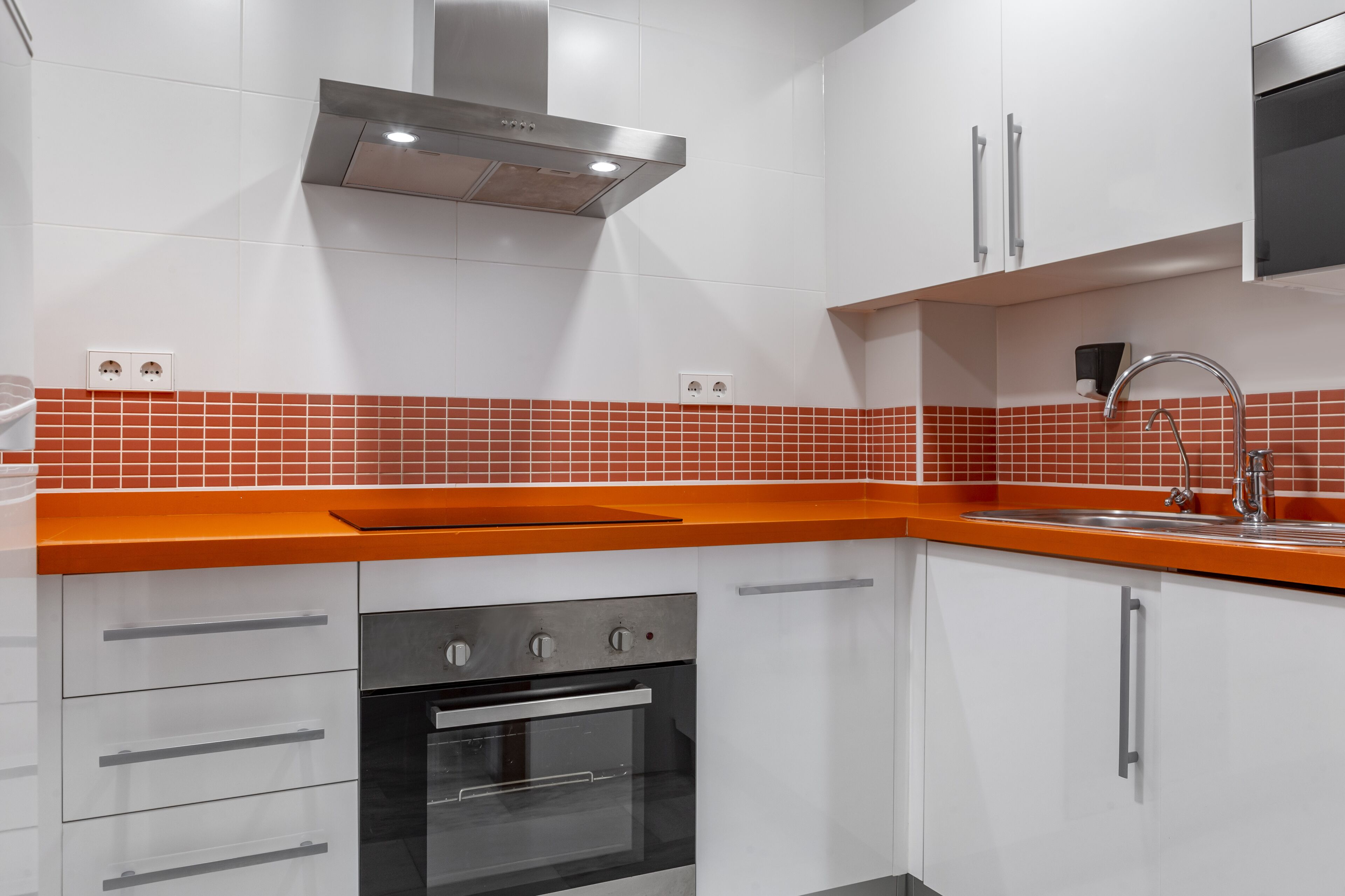 A modern kitchen with white cabinets, a stainless steel range hood, a red-tiled backsplash, and a built-in oven.