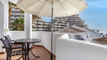 Een balkon met een tafel, stoelen, een grote parasol en uitzicht op de zee en gebouwen.