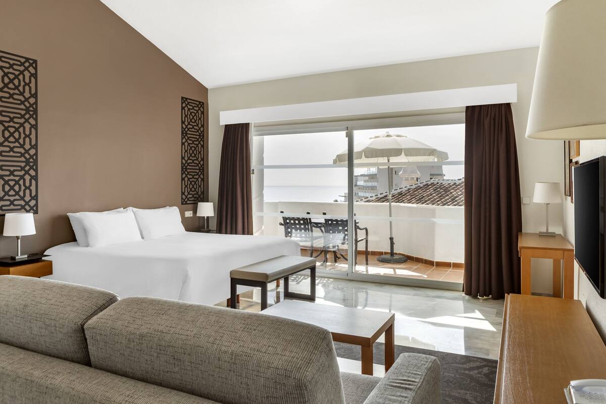 Une chambre d’hôtel avec un lit, un canapé, une table, une télévision et un balcon donnant sur la ville.