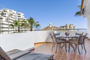 Una terraza con mesa lista para comer, rodeada de sillas y vista a un paisaje urbano con edificios y palmeras.