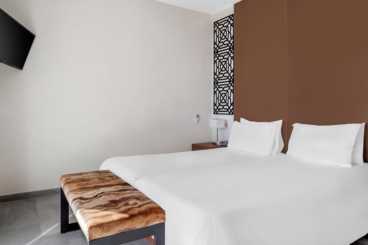 Moderni hotellihuone, jossa on suuri sänky, puinen penkki, seinälle asennettu televisio ja koristeellinen paneeli.