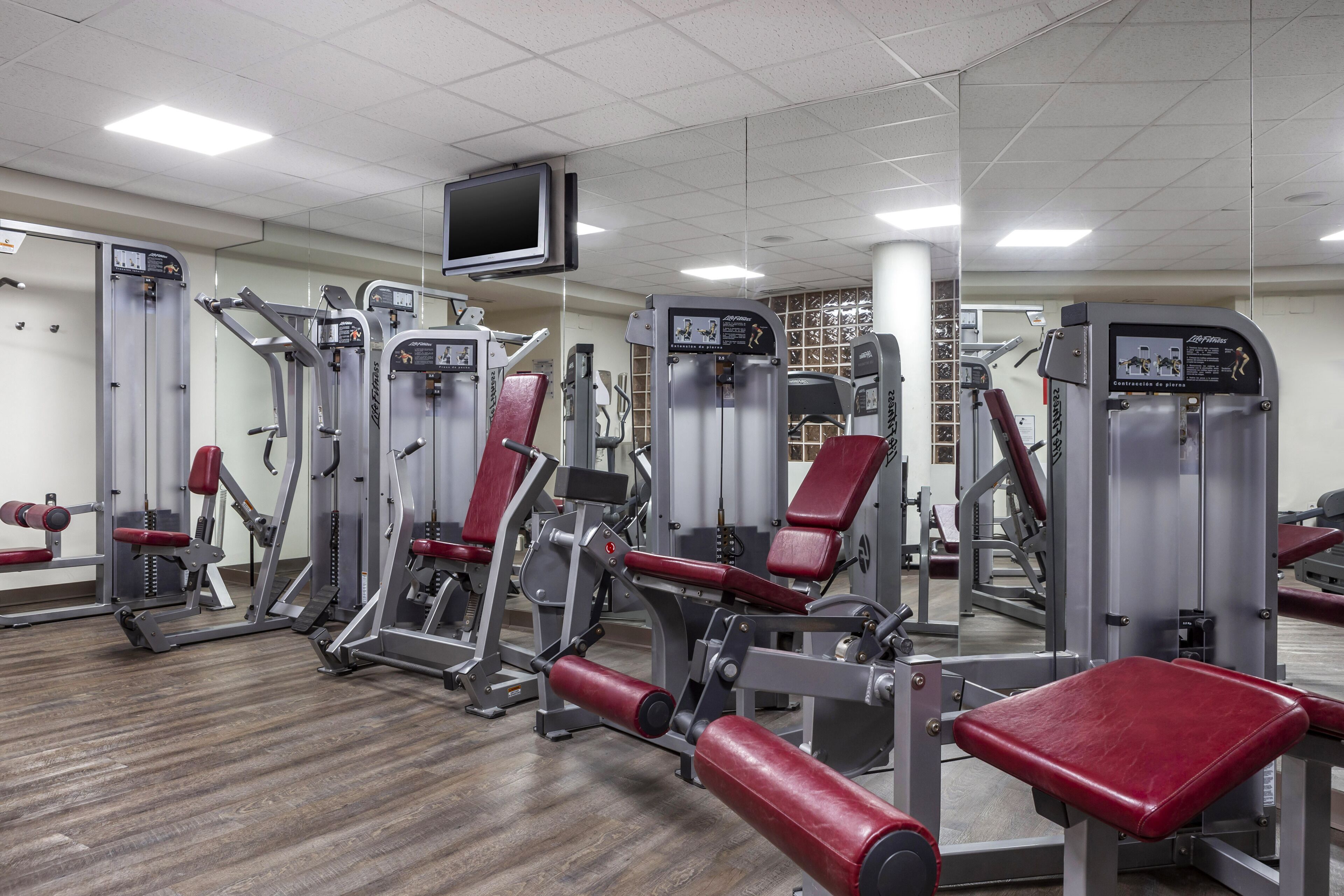 Un gimnasio con diversas máquinas de ejercicio, un televisor montado en la pared y suelo de madera.