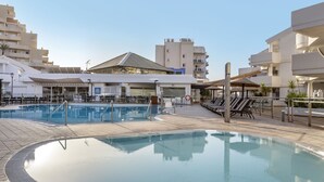 A swimming pool area with lounge chairs and a covered seating area, surrounded by modern buildings. - Sahara Sunset (Benalmádena)