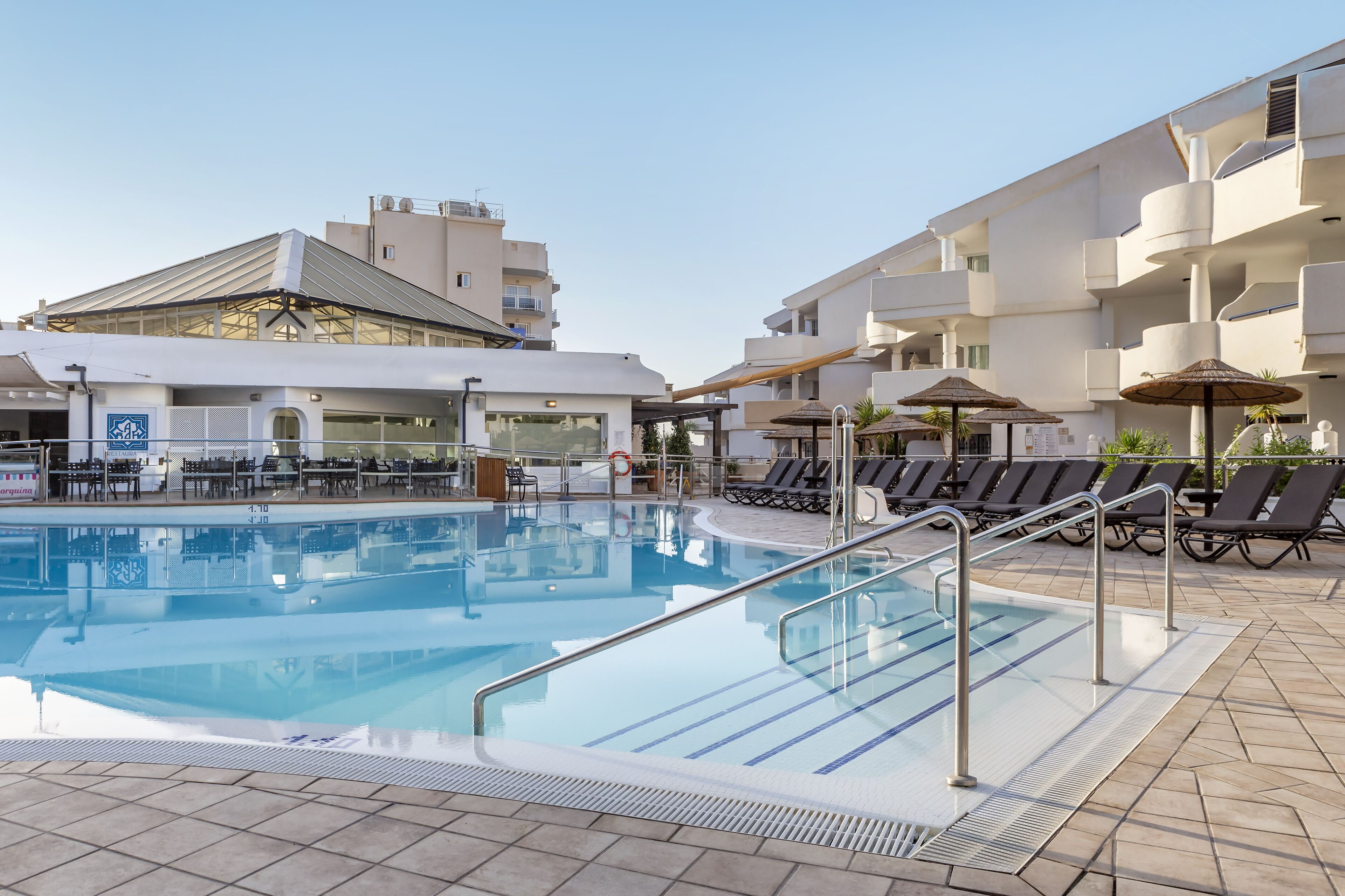 A swimming pool with sun loungers and umbrellas, surrounded by a building with balconies and a covered area with tables and chairs.