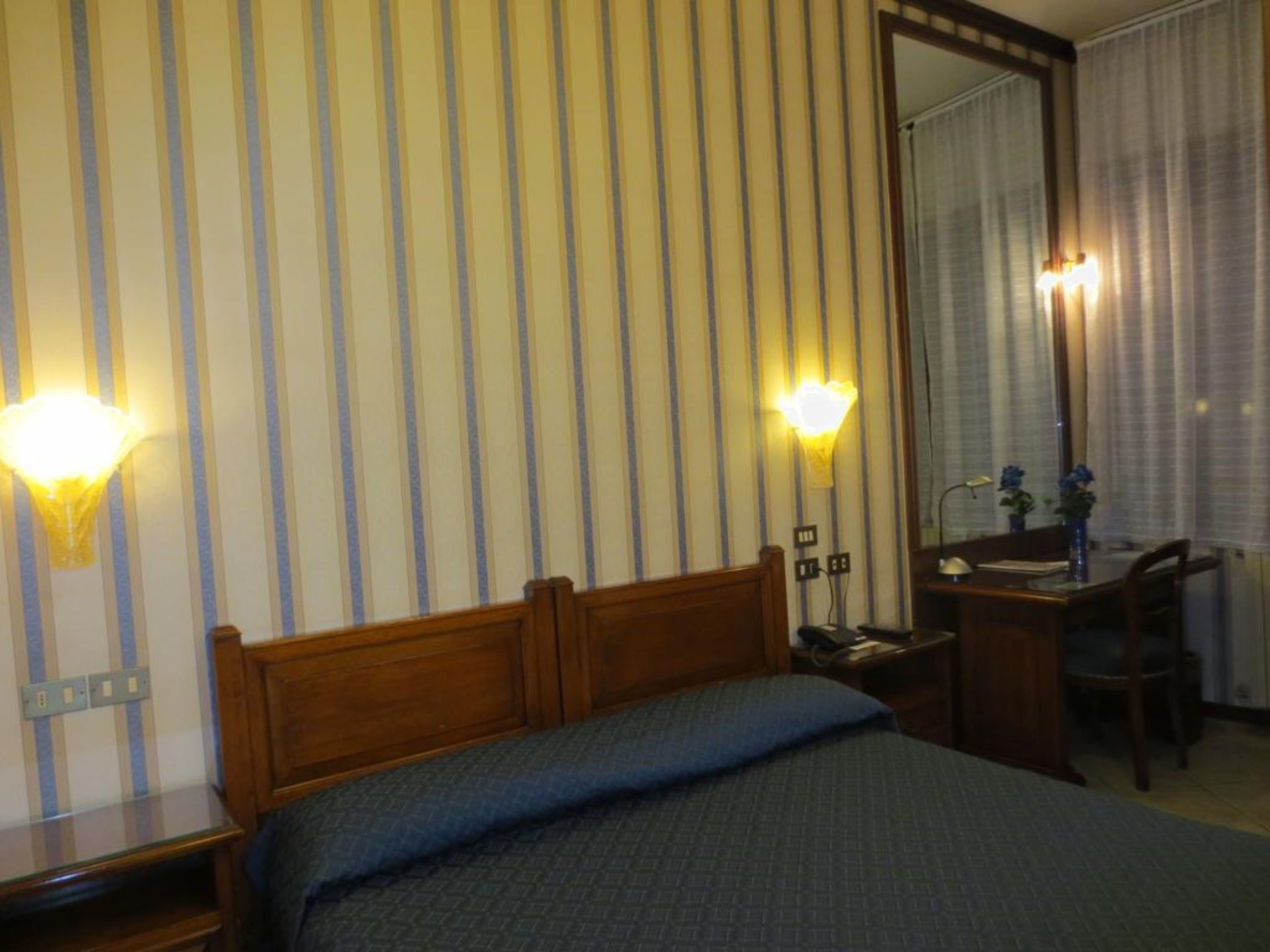 Habitación de hotel con cama, escritorio, silla y un espejo grande. Hay dos lámparas de pared y una ventana con cortinas.