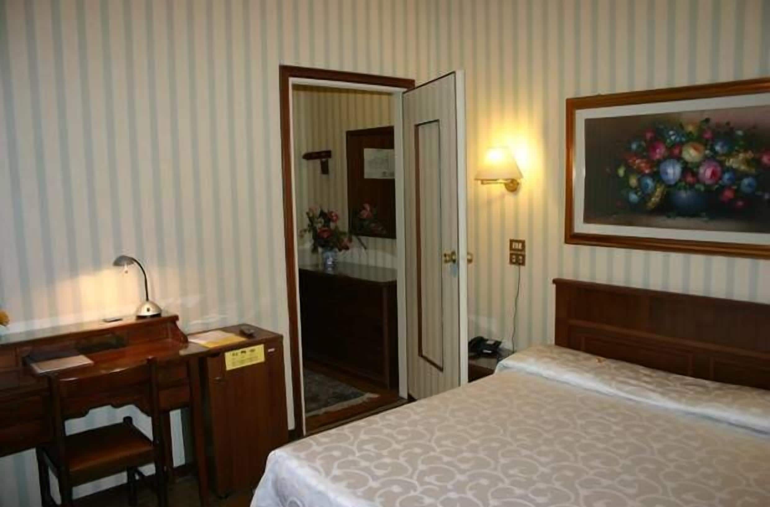 A hotel room with a bed, a desk, a chair, a nightstand, a lamp, a painting, and a door leading to a bathroom.