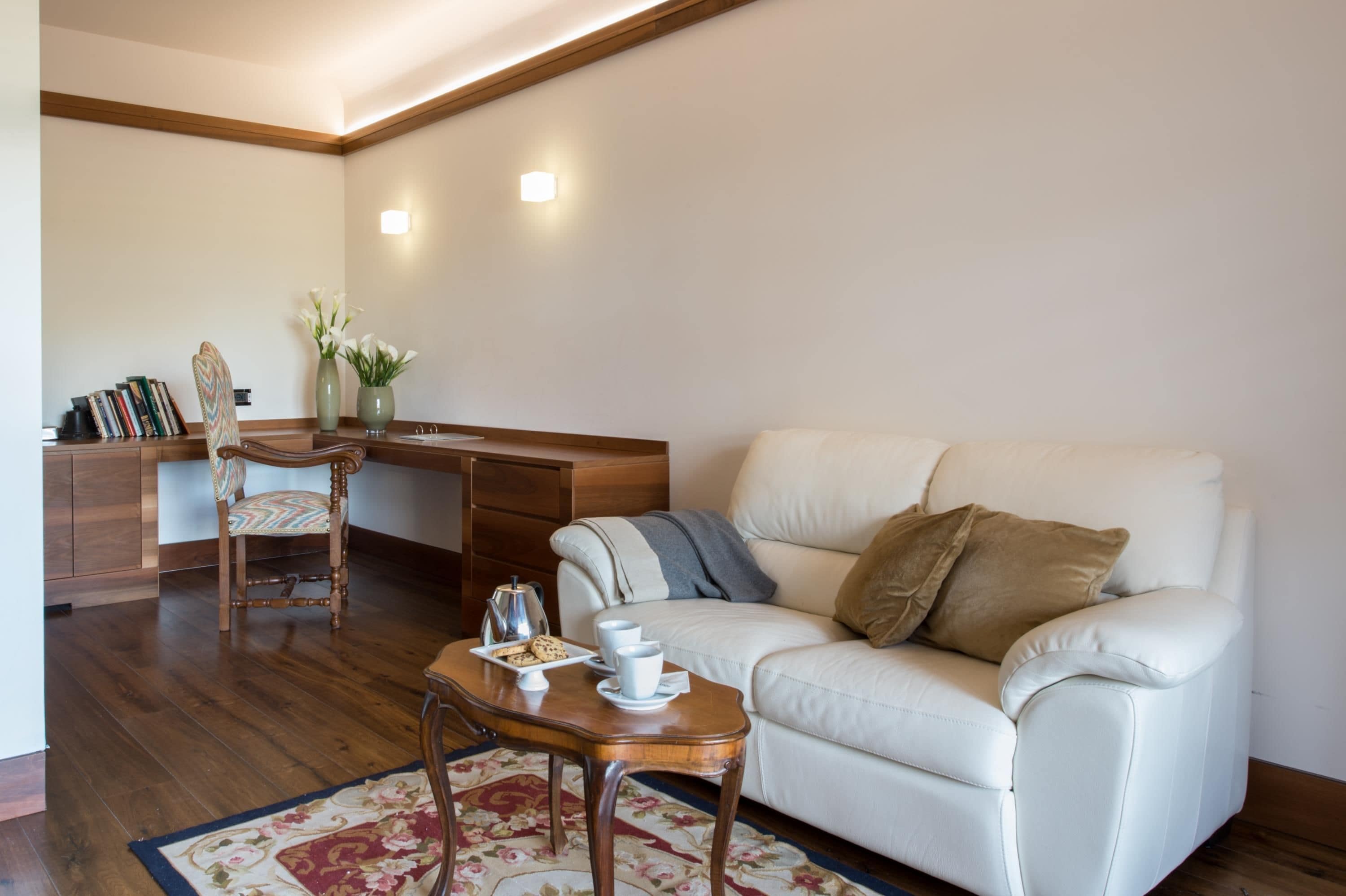 A modern living room with a white leather sofa, a wooden coffee table, a wooden desk with a chair, and a bookshelf.