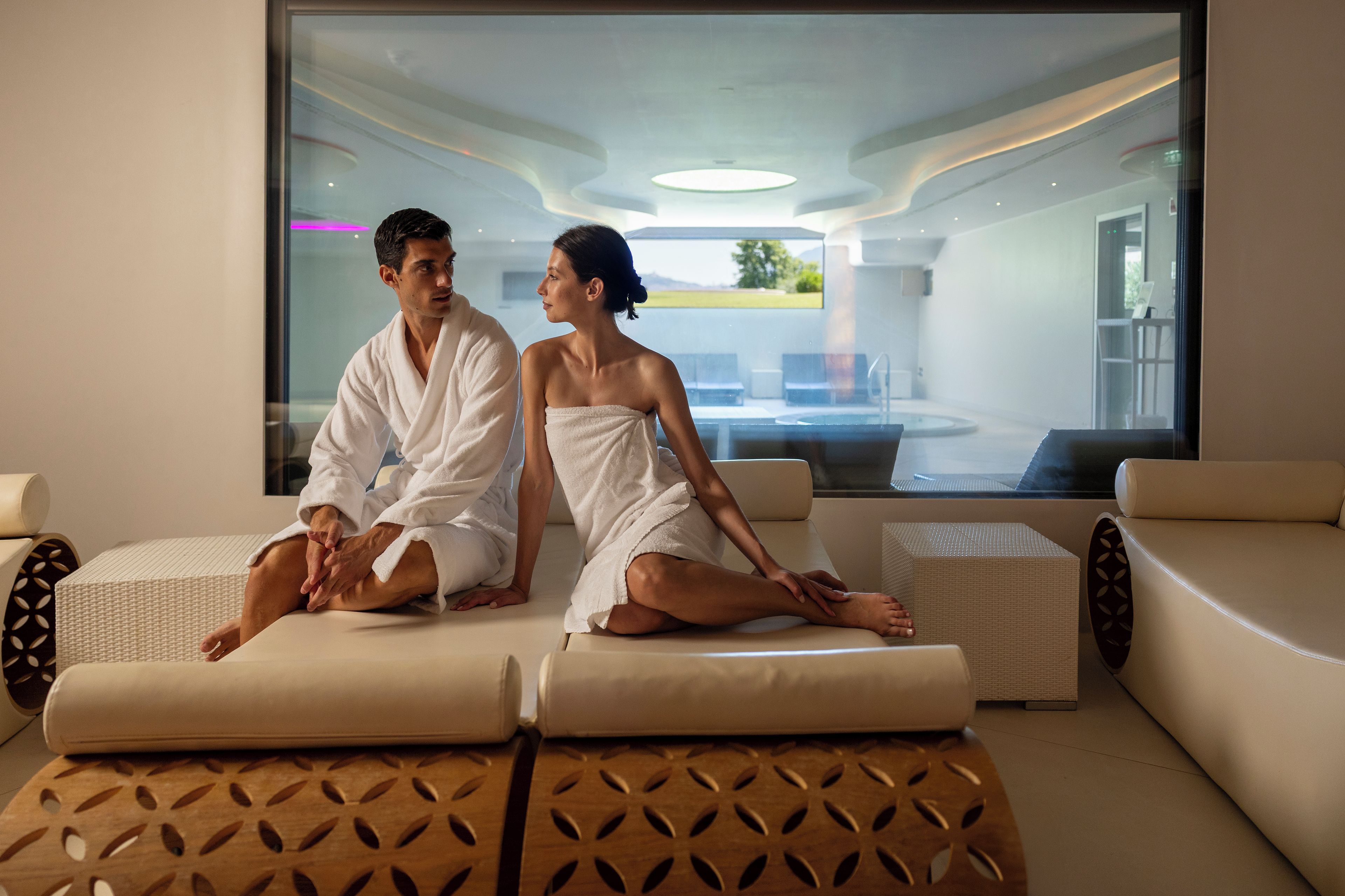 Un couple est assis sur une table de massage dans une salle de spa, avec vue sur une piscine et un espace extérieur.