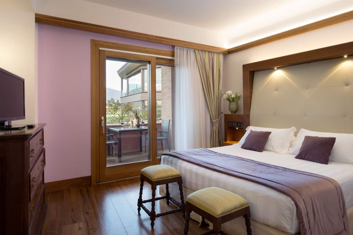 A hotel room with a large bed, a wooden dresser, a television, a balcony with a table and chairs, and a view of the outside.
