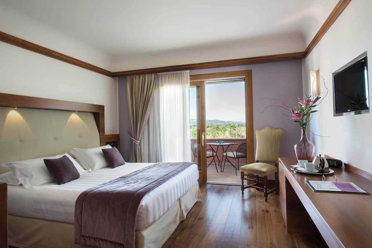 A hotel room with a large bed, a desk with a chair, a TV, and a balcony with a table and chairs.