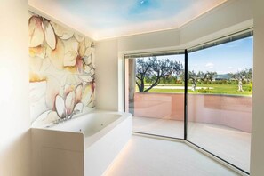 Un baño moderno con una bañera grande, un lavamanos empotrado en la pared y un amplio ventanal de vidrio que da a un balcón con vistas a árboles y un área verde.