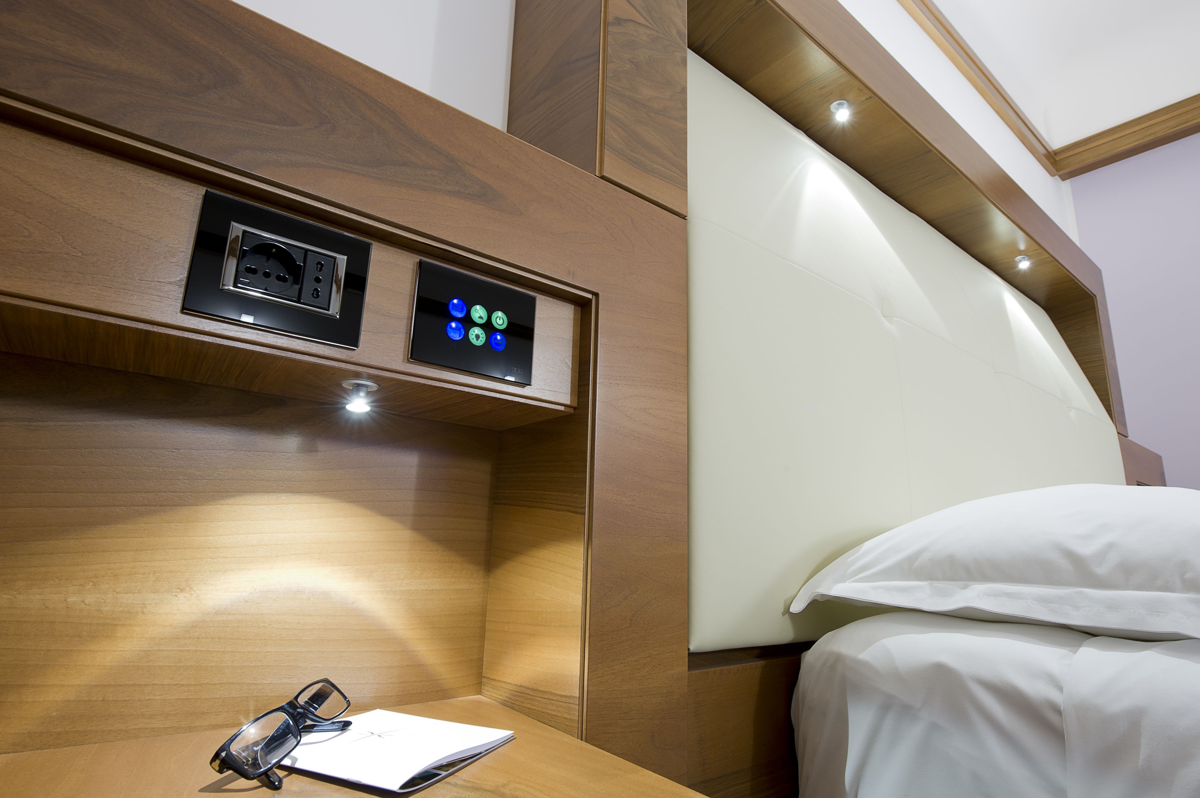 A hotel room with a wooden headboard, a bedside table with glasses and a notebook, and a control panel above the bed.