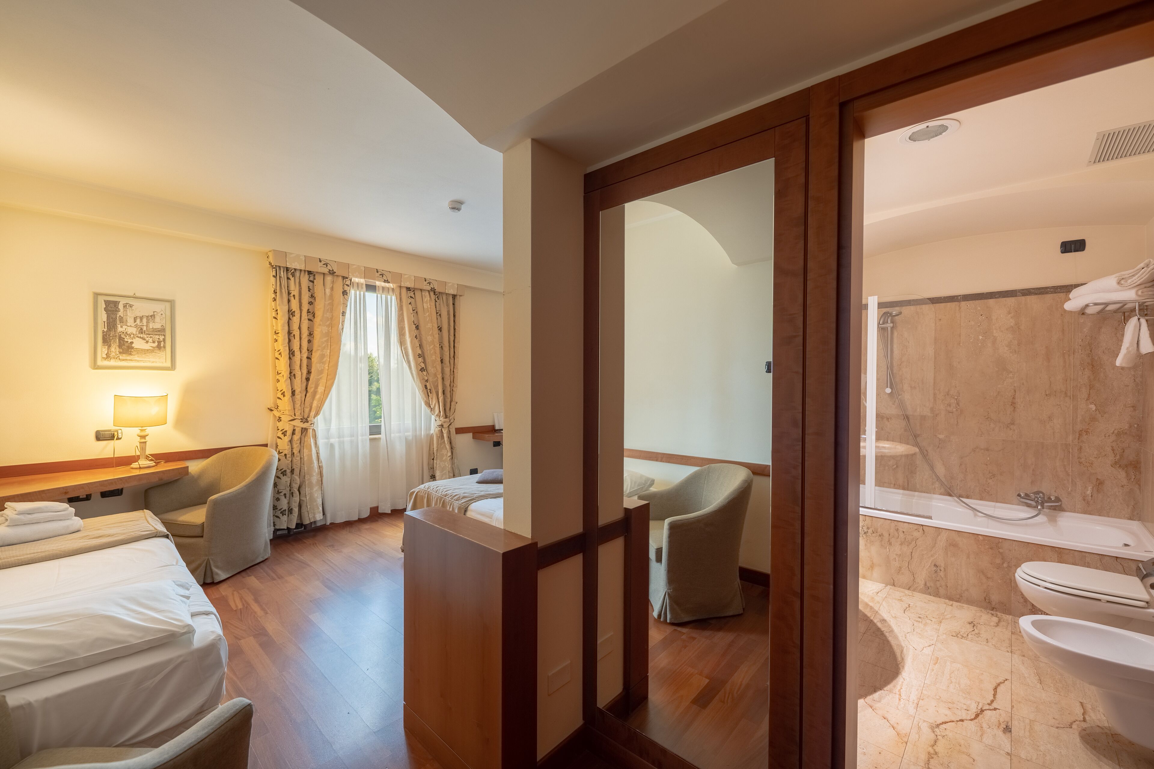 A hotel room with a bed, two chairs, a desk with a lamp, a window with curtains, and a view into a bathroom with a shower and toilet.