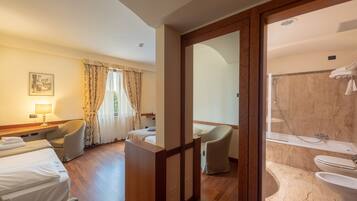A hotel room with a bed, two chairs, a desk with a lamp, a window with curtains, and a view into a bathroom with a shower and toilet.