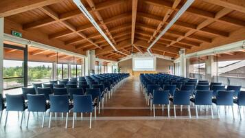 Ein geräumiger Saal mit blauen Stuhlreihen, einer hohen Holzbalkendecke und großen Fenstern mit Blick auf Grünflächen.