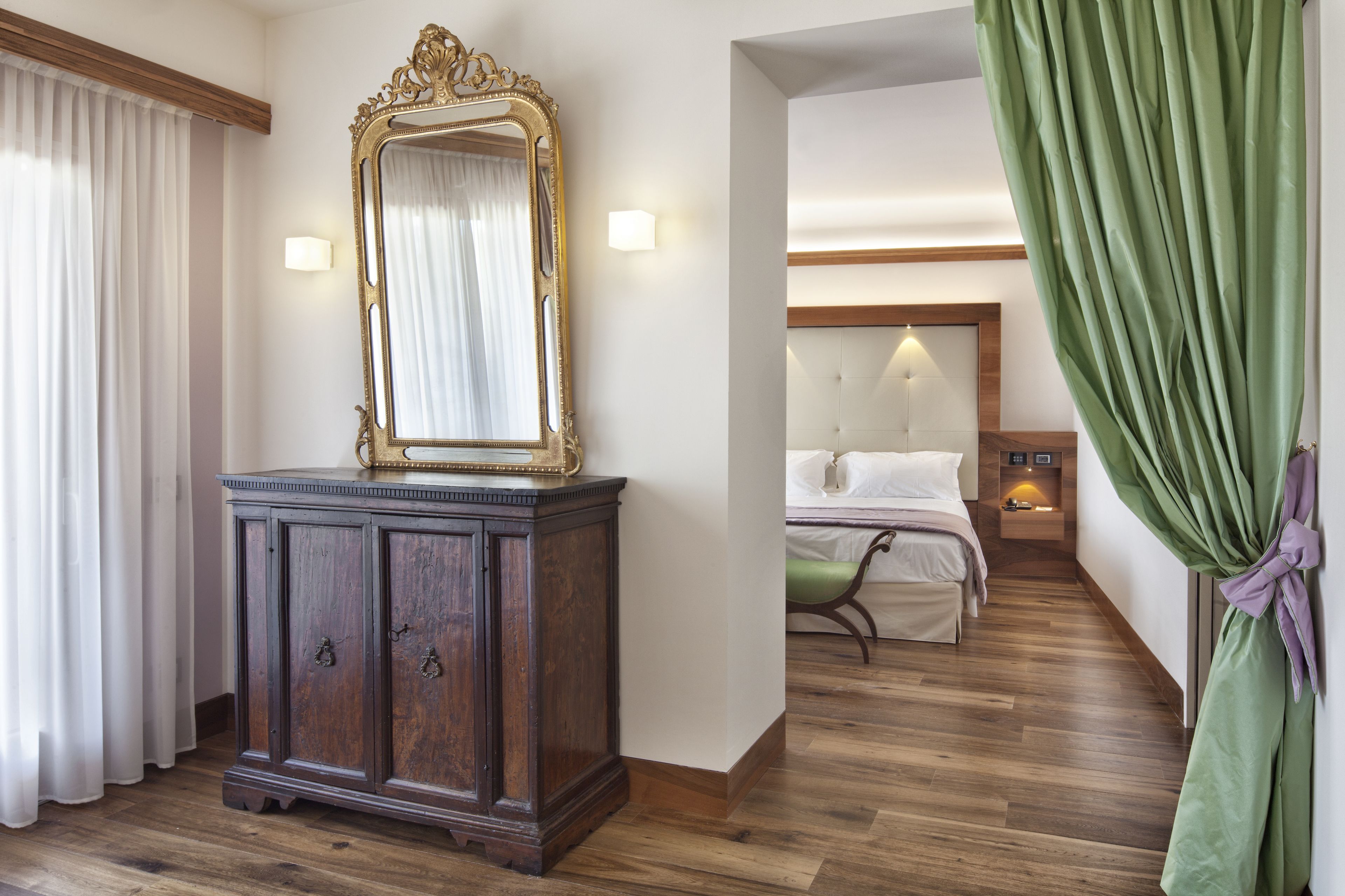 Ein Hotelzimmer mit Holzboden, einem großen Spiegel, einem Nachttisch, einem Bett mit weißer Bettwäsche und einem grünen Vorhang.