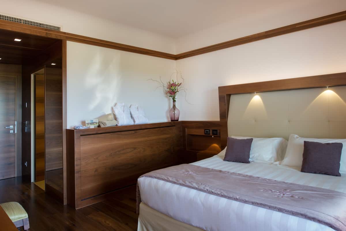 A modern hotel room with a wooden headboard, a large bed with white and brown bedding, a bedside table with a vase of flowers, and a wooden wardrobe.