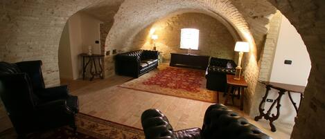 An arched room with a fireplace, leather armchairs, a red carpet, and a wooden table with a lamp.