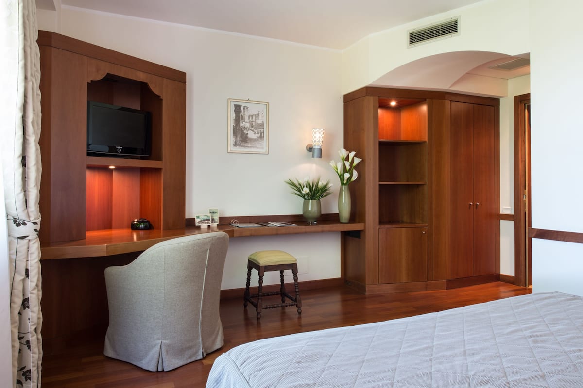 A hotel room with a bed, a desk with a television, a chair, a small stool, a vase with flowers, and wooden wardrobes.
