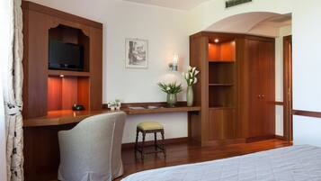 A hotel room with a bed, a desk with a television, a chair, a small stool, a vase with flowers, and wooden wardrobes.