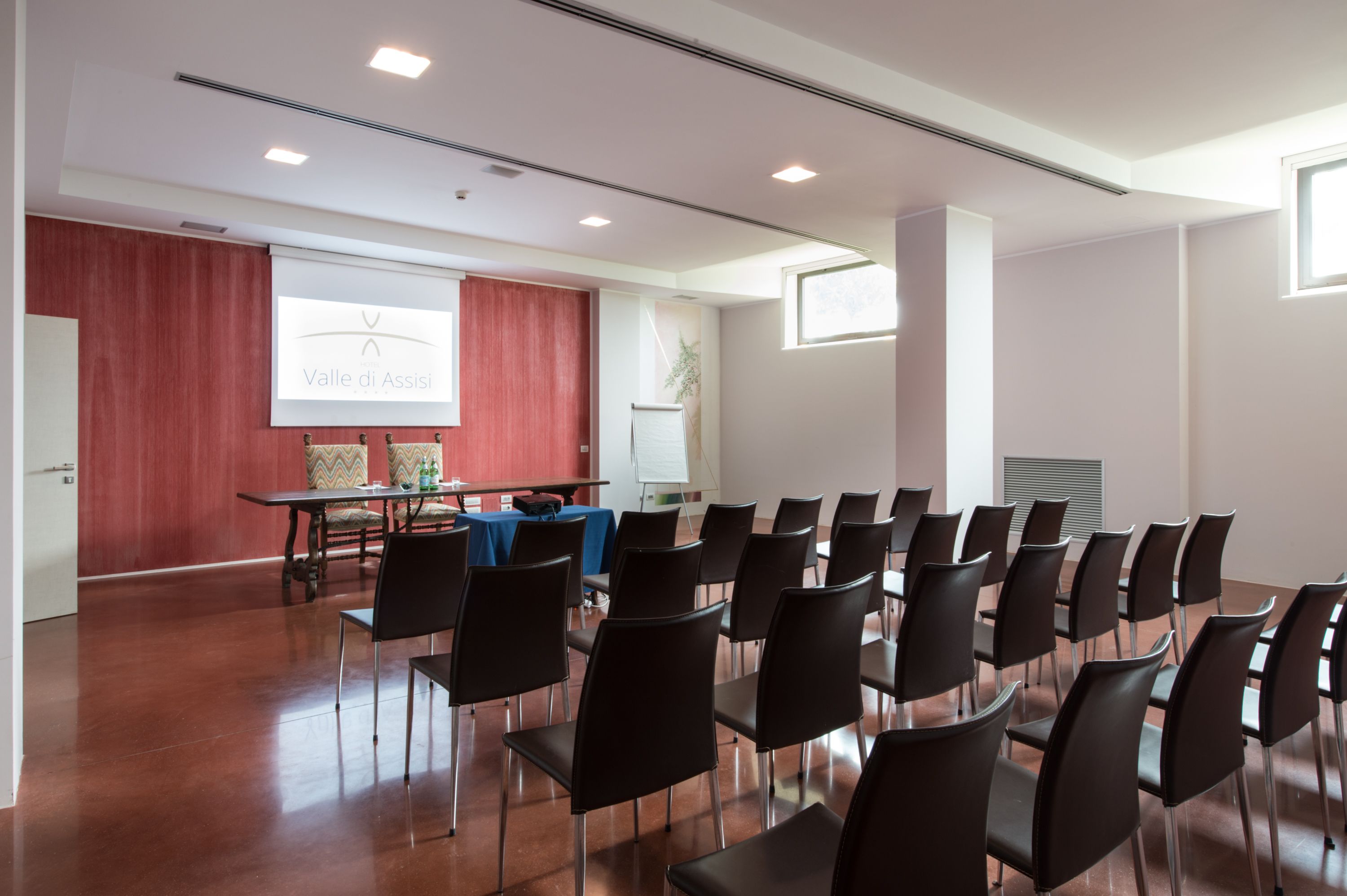 Une salle de conférence avec des rangées de chaises, un écran de présentation et une table recouverte d’une nappe bleue.