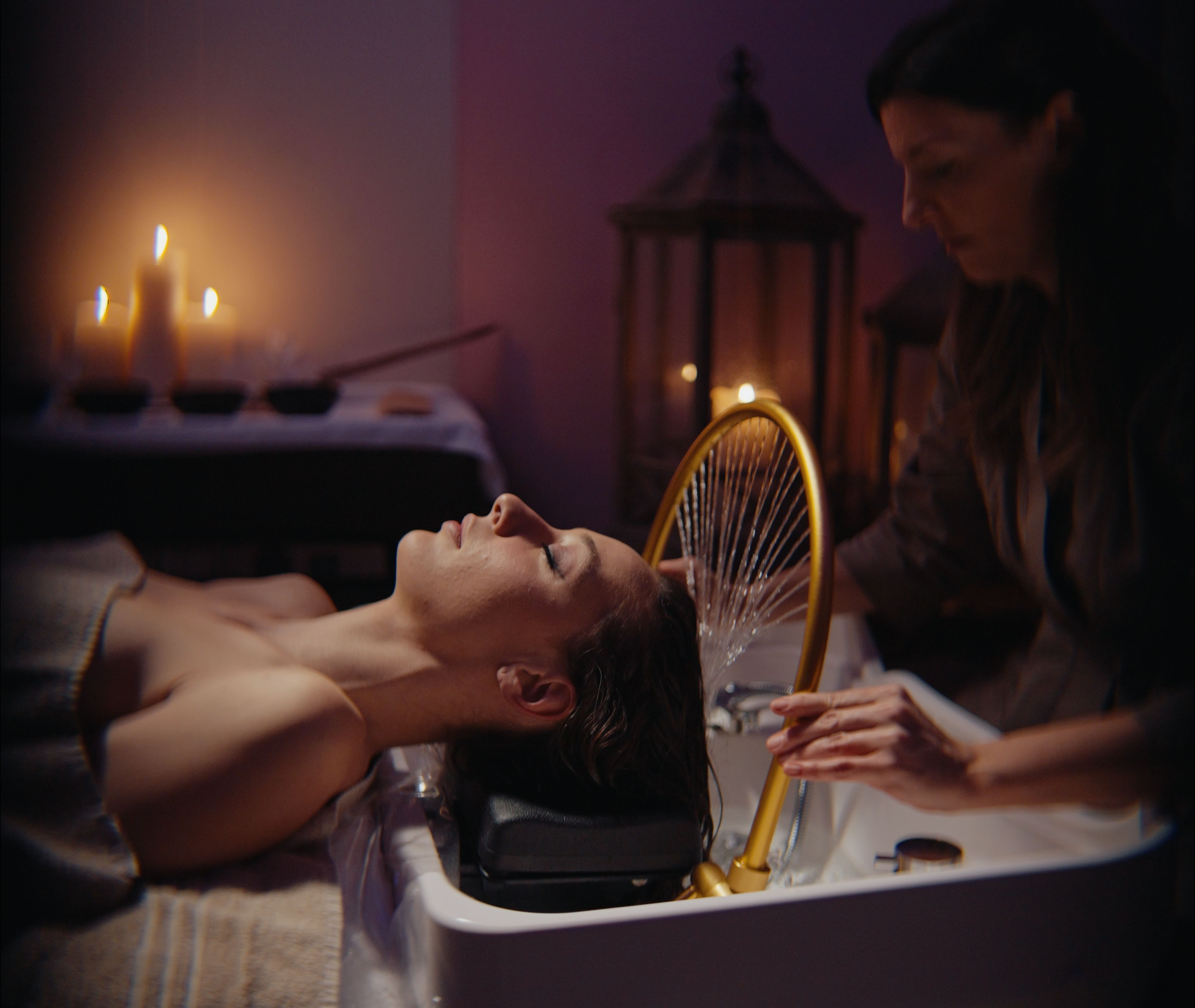 A person lying in a bathtub with a headrest, another person attending to them, and candles in the background.