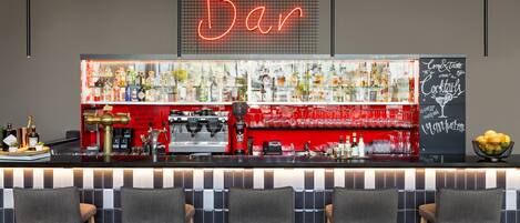 A bar area with a neon sign, a variety of bottles, a coffee machine, and bar stools.