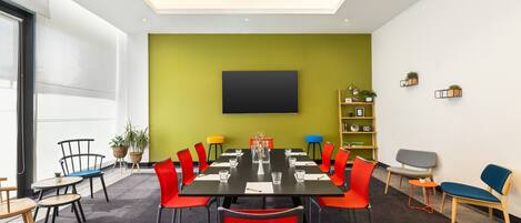 A conference room with a large table, red and black chairs, a green accent wall, and a mounted TV.