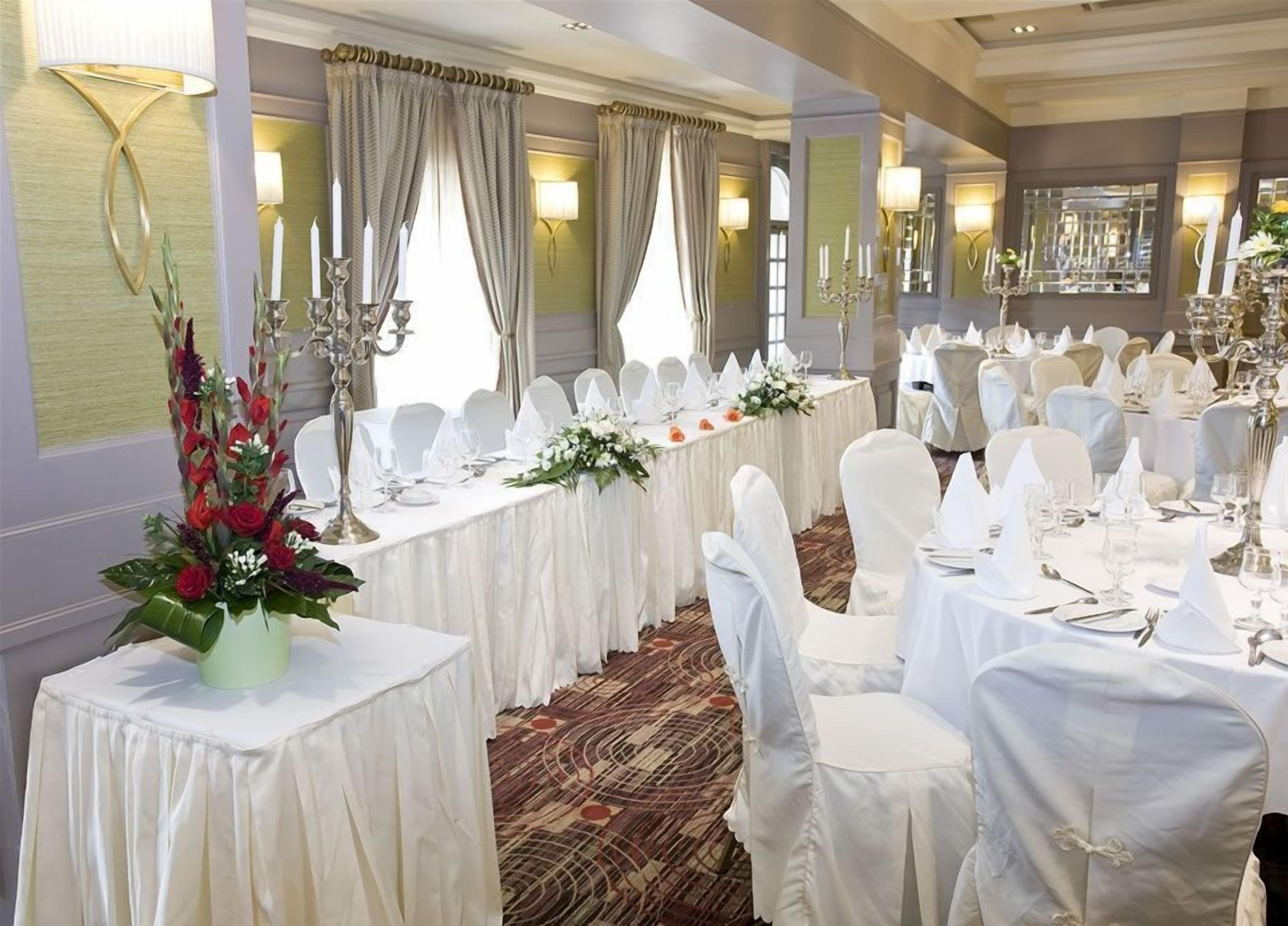 Une salle de banquet avec des tables rondes dressées pour un événement formel, agrémentée de nappes blanches, de chaises recouvertes de housses blanches et de compositions florales en guise de centre de table.