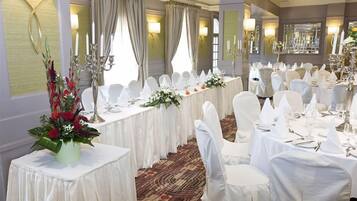A banquet hall with round tables set for a formal event, featuring white tablecloths, chairs with white covers, and floral centerpieces.