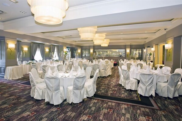 Un salón de banquetes con mesas redondas cubiertas con manteles blancos y sillas, grandes candelabros y suelo alfombrado.