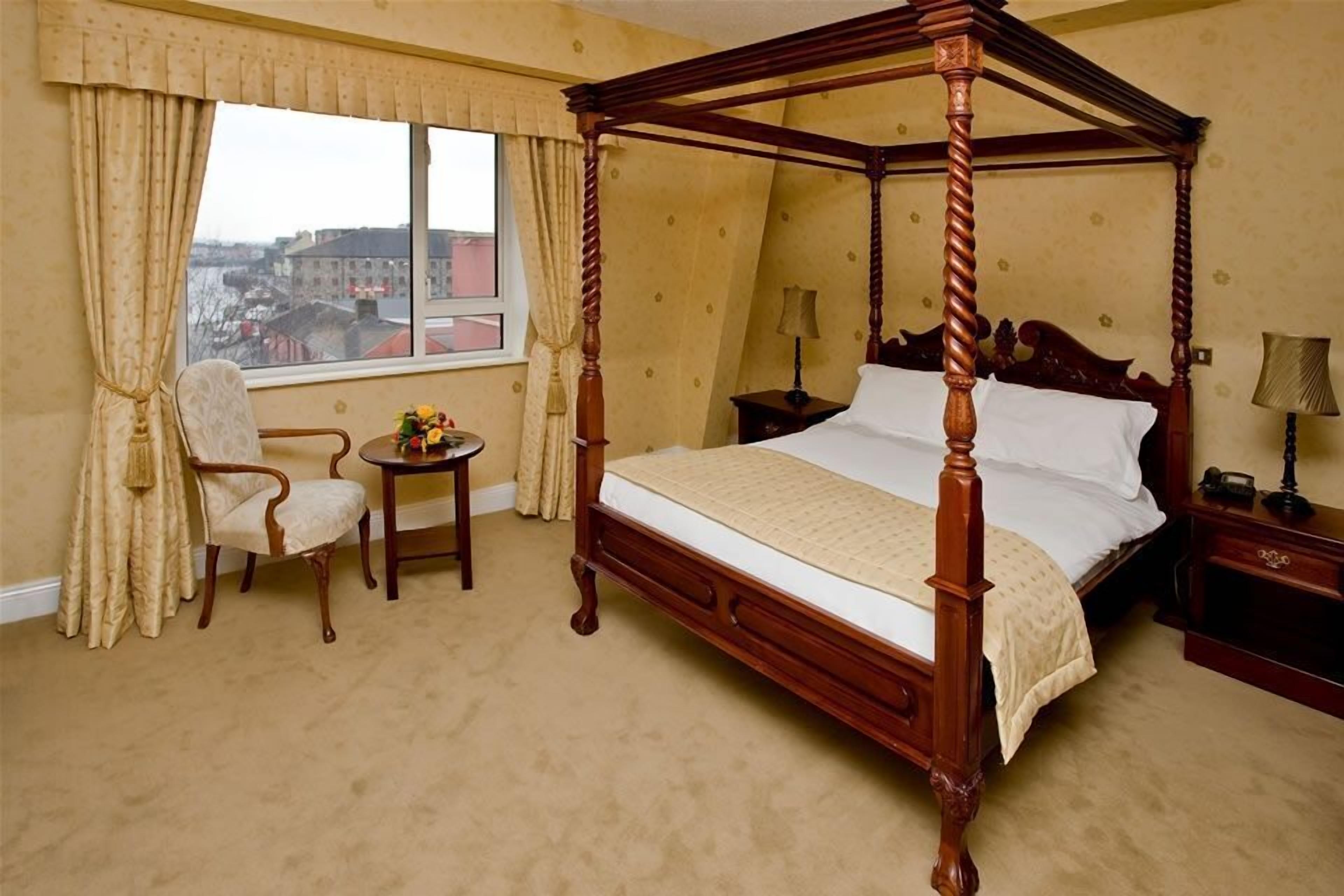 Una cama con dosel de madera, una mesita redonda con una lámpara, una silla y vistas a edificios a través de una ventana con cortinas.